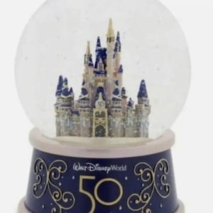 Peut inclure: Un globe de neige avec un château miniature détaillé aux accents bleus et dorés. La base bleu marine porte des motifs dorés et le texte "Walt Disney World 50". Le globe est transparent et rempli de paillettes.