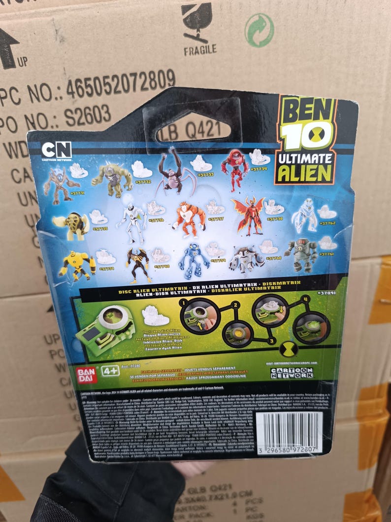 Op de afbeelding: Achterkant van een Ben 10 Ultimate Alien speelgoedverpakking. De verpakking bevat verschillende illustraties van buitenaardse personages en het Cartoon Network-logo. De tekst "DISC BLIEN ULTIMATRIN" en "4+" is inbegrepen.