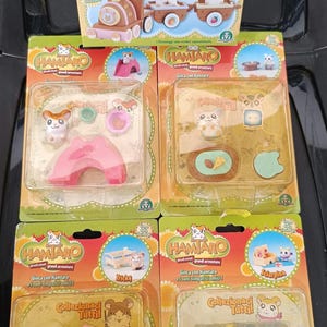 Op de afbeelding: Een verzameling Hamtaro-speelgoedsets, met verschillende Hamtaro-personages en accessoires. De sets bevatten een trein, een roze boog, een miniatuur tafelsetting, een winkelwagentje en een piano. De verpakking bevat de tekst "Il Trenino Ciuf Ciuf".