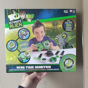 Könnte beinhalten: Ein Ben 10 Ultimate Alien Hero Time Omnitrix Spielzeugset in einer rechteckigen Schachtel. Die Verpackung zeigt ein Kind, das grüne und schwarze Uhrenkomponenten zusammenbaut, mit Text, der 35 Teile und über 1000 Kombinationen angibt. Die Schachtel enthält Grafiken des Omnitrix, eine Alien-Figur und den Text Core Lights Up.