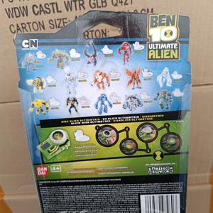 May include: The back of a Ben 10 Ultimate Alien toy package. The package features various alien figures, the Cartoon Network logo, and the text "DISC BLIEN ULTIMETRIE - DE BLIEN ULTIMATRIN - DISKMATRIN". The package also includes the Bandai logo.