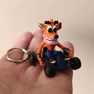 May include: A keychain featuring a small, orange Crash Bandicoot figurine driving a dark blue go-kart. The character has large green eyes, a wide grin with visible teeth, and black eyebrows. The go-kart has four black wheels and a metal ring.