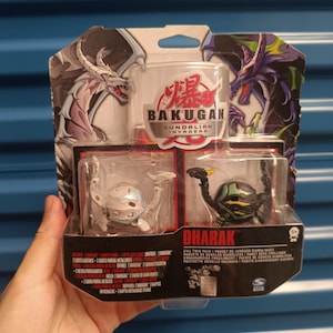 Peut inclure: Un coffret de jouets Bakugan Gundalian Invaders. L'emballage présente des images de dragons et le texte "BAKUGAN" et "DHARAK". L'ensemble comprend deux figurines Bakugan et des cartes. Les couleurs sont le noir, le vert, le blanc et le rouge.