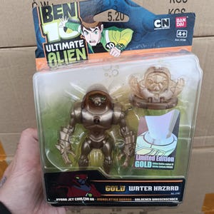 Op de afbeelding: Een goudkleurige Ben 10 Ultimate Alien actiefiguur in een plastic verpakking. De verpakking bevat een kleinere gouden figuur en de woorden "Limited Edition GOLD". De verpakking bevat ook het Ben 10-logo en de woorden "Gold Water Hazard".