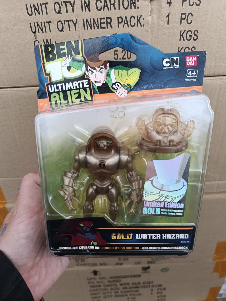 Op de afbeelding: Een goudkleurige Ben 10 Ultimate Alien actiefiguur in een plastic verpakking. De verpakking bevat een kleinere gouden figuur en de woorden "Limited Edition GOLD". De verpakking bevat ook het Ben 10-logo en de woorden "Gold Water Hazard".