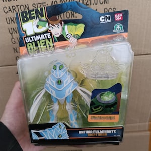 Op de afbeelding: Een Ben 10 Ultimate Alien Amphibian actiefiguur in een transparante plastic verpakking. De figuur is lichtblauw en wit, met een uniek ontwerp. De verpakking bevat de logo's van Cartoon Network en Bandai.