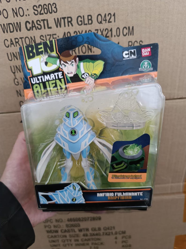 Op de afbeelding: Een Ben 10 Ultimate Alien Amphibian actiefiguur in een transparante plastic verpakking. De figuur is lichtblauw en wit, met een uniek ontwerp. De verpakking bevat de logo's van Cartoon Network en Bandai.
