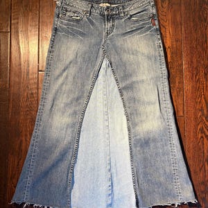 Può includere: Una gonna di jeans lavata chiara con un design a spacco frontale. La gonna ha un classico design a cinque tasche, chiusura con bottone e cerniera e orlo sfilacciato. Il denim presenta sottili dettagli di sbiaditura e baffi.