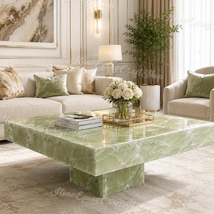 May include: A square, light green onyx coffee table with a pedestal base. The table has a polished surface and is in a living room setting with a sofa, armchair, and decorative items. Two books are on the table.
