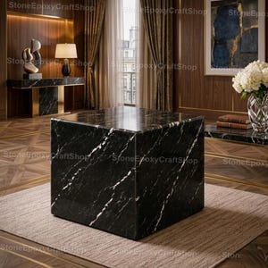 May include: A large, square black marble coffee table with white veining. The table is placed on a neutral-colored rug in a room with wood paneling and gold accents. The room also features a matching marble console table.