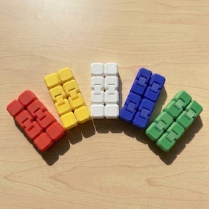 Könnte beinhalten: Eine Sammlung farbenfroher Fidget-Spielzeuge in Rot, Gelb, Weiß, Blau und Grün. Jedes Spielzeug besteht aus verbundenen, ineinandergreifenden Blöcken. Die Spielzeuge sind auf einer hellbraunen Holzoberfläche angeordnet, mit Schatten.