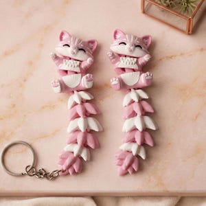 May include: Two pink and white cat-shaped fidget toys with a keychain. The cats have their arms up and are connected to a series of leaf-shaped segments. The toys are on a pink marble surface.