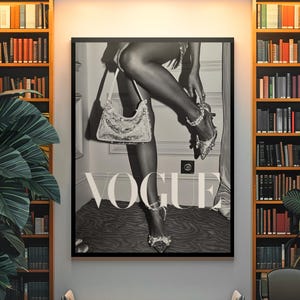 May include: Black and white Vogue poster featuring a woman adjusting a jeweled shoe, holding a sequined purse. The poster is framed and displayed in a room with bookshelves, a bench, and a chair.