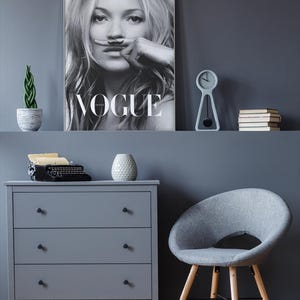 May include: An interior scene with a grey chest of drawers, a grey chair with wooden legs, and a framed Vogue magazine cover. A small clock, books, and a potted plant are on a shelf above the chest of drawers.