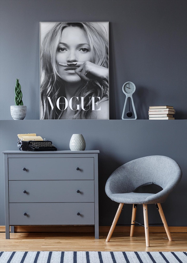 May include: An interior scene with a grey chest of drawers, a grey chair with wooden legs, and a framed Vogue magazine cover. A small clock, books, and a potted plant are on a shelf above the chest of drawers.