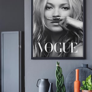 May include: Monochrome framed Vogue magazine cover with a woman and a finger mustache. The image is above a grey cabinet with decorative items, including a green plant, vases, and an orange bottle.
