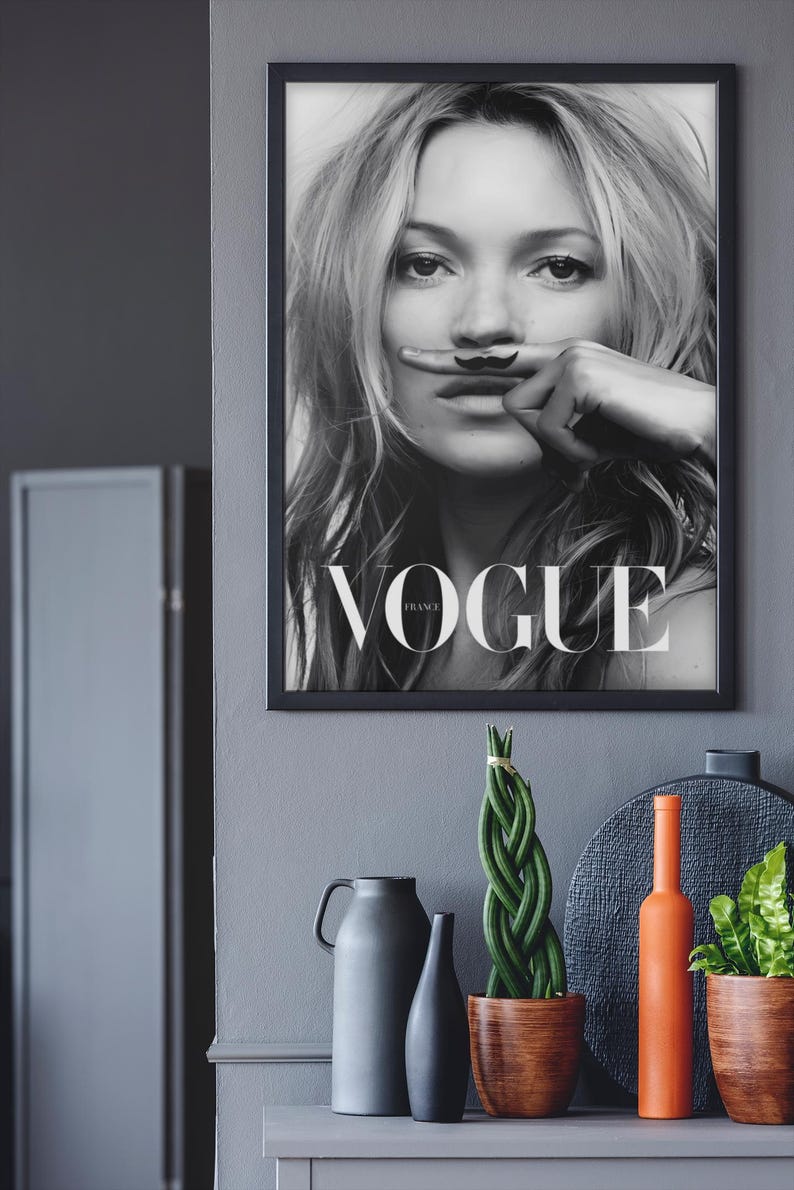 May include: Monochrome framed Vogue magazine cover with a woman and a finger mustache. The image is above a grey cabinet with decorative items, including a green plant, vases, and an orange bottle.