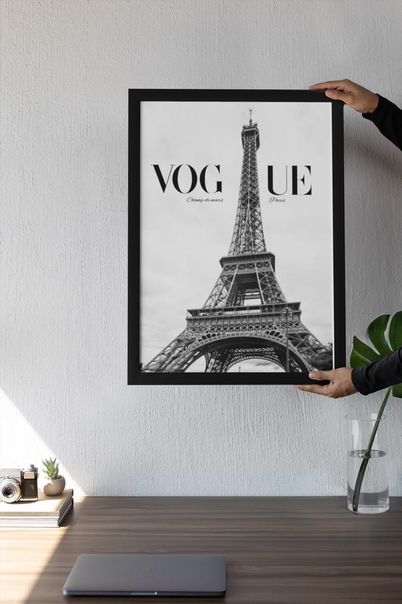 May include: Monochrome framed print of the Eiffel Tower. The print displays the word "VOGUE" at the top, with "Champ de mars" and "Paris" below. The image is held up by a person.
