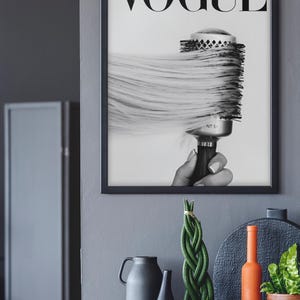 May include: Black and white framed print featuring the word "VOGUE" at the top. The image shows a round hairbrush with long, flowing hair. Decorative items, including vases and plants, are on a gray surface below the print.