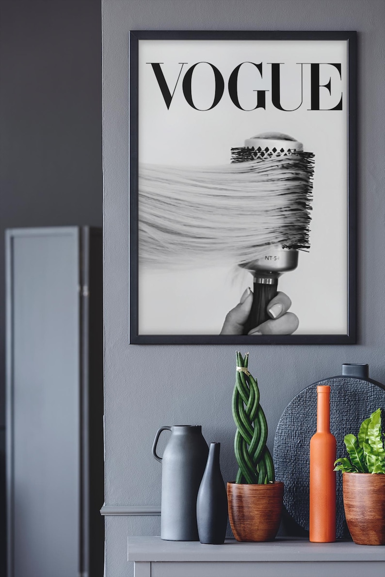 May include: Black and white framed print featuring the word "VOGUE" at the top. The image shows a round hairbrush with long, flowing hair. Decorative items, including vases and plants, are on a gray surface below the print.