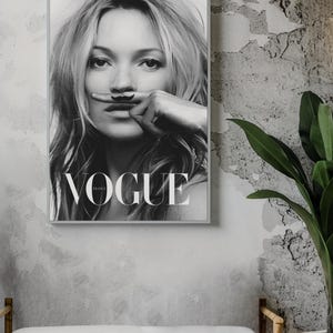 May include: Black and white framed Vogue France magazine cover featuring a woman with a finger moustache. The magazine cover is displayed on a grey wall above a bench with a white cushion. A potted plant is to the right.