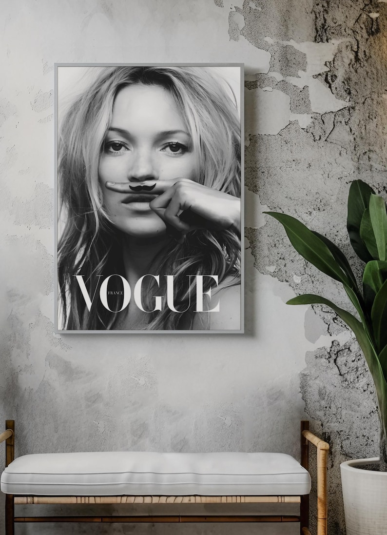 May include: Black and white framed Vogue France magazine cover featuring a woman with a finger moustache. The magazine cover is displayed on a grey wall above a bench with a white cushion. A potted plant is to the right.