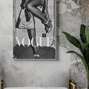 May include: Black and white framed Vogue print. The image features a person wearing high heels and holding a handbag. The word "VOGUE" is prominently displayed in large letters. The print is displayed on a textured gray wall.