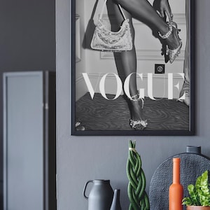 May include: Black and white framed Vogue magazine cover featuring a woman with a sequined handbag and high heels. Decorative items include a gray vase, a black vase, a braided plant in a wooden pot, and an orange bottle.