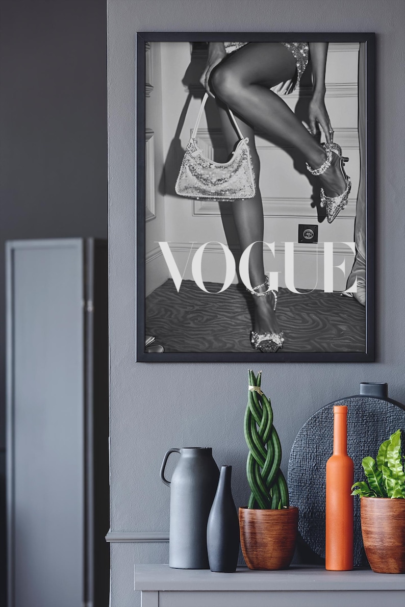 May include: Black and white framed Vogue magazine cover featuring a woman with a sequined handbag and high heels. Decorative items include a gray vase, a black vase, a braided plant in a wooden pot, and an orange bottle.