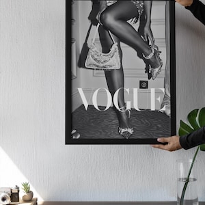 May include: Black and white framed Vogue print featuring a woman's legs, high heels, and a sequined handbag. The image has the Vogue logo in large letters. The print is held by a person.