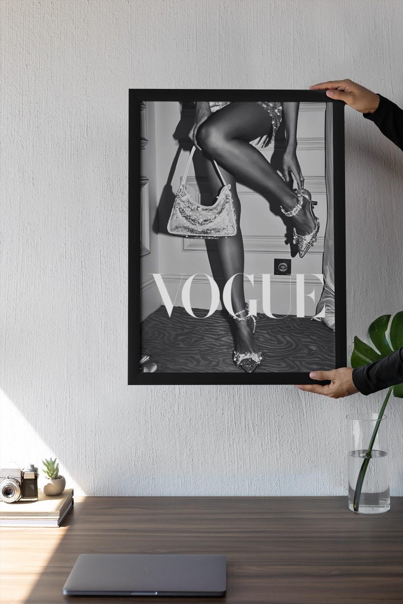 May include: Black and white framed Vogue print featuring a woman's legs, high heels, and a sequined handbag. The image has the Vogue logo in large letters. The print is held by a person.