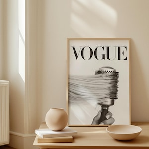 May include: A framed black and white Vogue print featuring a hairbrush with long hair. The print is displayed on a light wood table with a small beige vase, books, and a bowl. The room has a neutral color palette.
