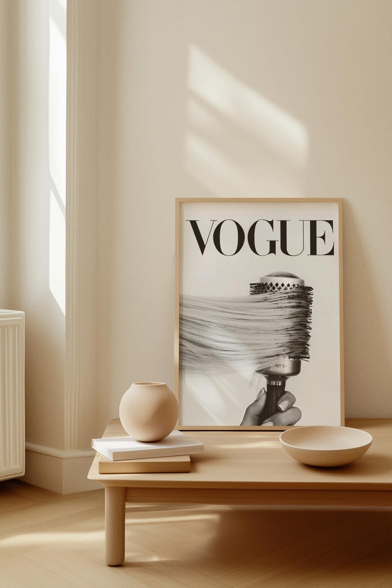 May include: A framed black and white Vogue print featuring a hairbrush with long hair. The print is displayed on a light wood table with a small beige vase, books, and a bowl. The room has a neutral color palette.
