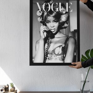 Może przedstawiać: Czarno-biały oprawiony wydruk przedstawiający kobietę z wałkami do włos&oacute;w, trzymającą telefon. Widoczne logo Vogue. Wydruk trzymany przez osobę. Estetyka vintage.