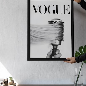 May include: Black and white framed print featuring the word "VOGUE" at the top. The image shows a round hairbrush with long, flowing hair. The print is held by a person.