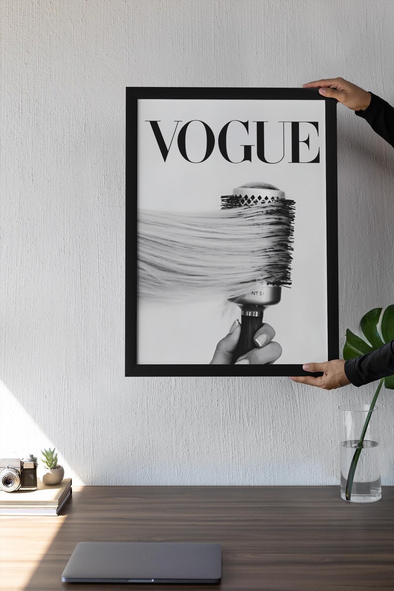 May include: Black and white framed print featuring the word "VOGUE" at the top. The image shows a round hairbrush with long, flowing hair. The print is held by a person.