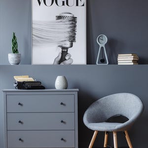 May include: A gray dresser with three drawers, a gray chair, and a Vogue poster. The poster features a black and white image of a hairbrush. A small clock, books, and a plant are on a shelf above the dresser.