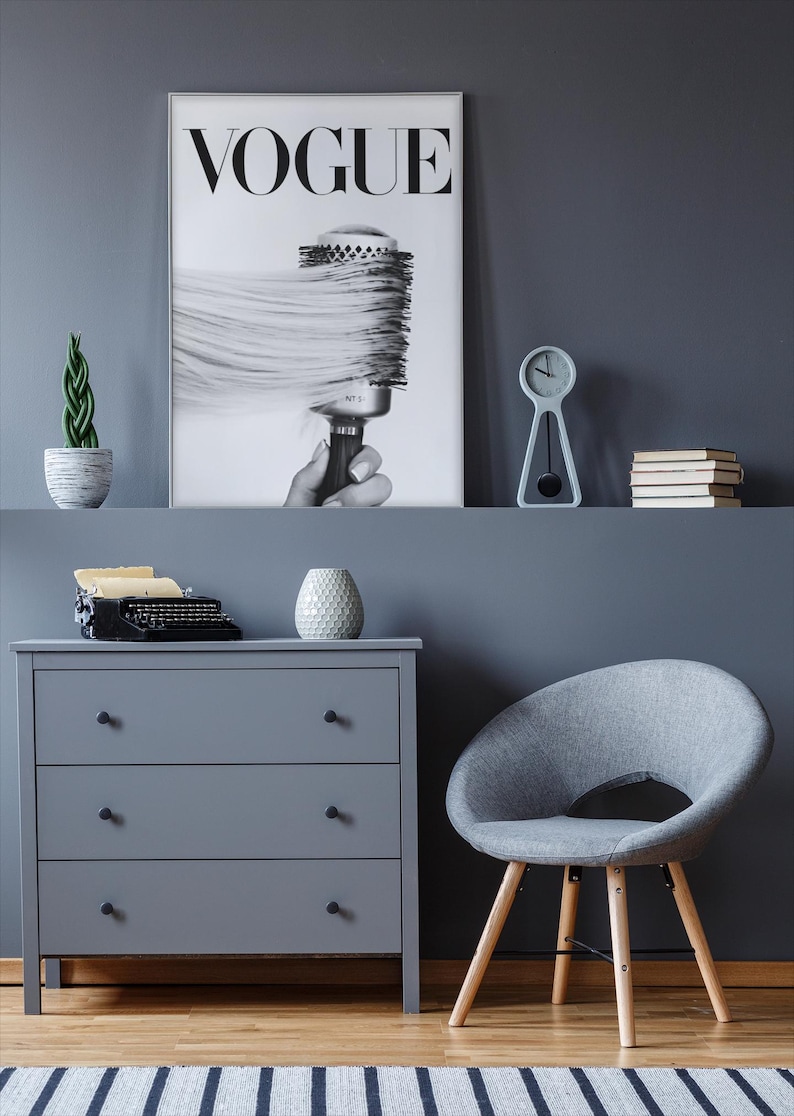 May include: A gray dresser with three drawers, a gray chair, and a Vogue poster. The poster features a black and white image of a hairbrush. A small clock, books, and a plant are on a shelf above the dresser.