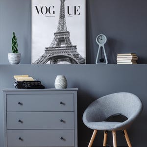 May include: A stylish grey room featuring a Vogue Paris print with the Eiffel Tower. A grey chest of drawers, a grey chair, a small clock, and a potted plant are also present. A vintage typewriter and a decorative vase sit on the chest of drawers.