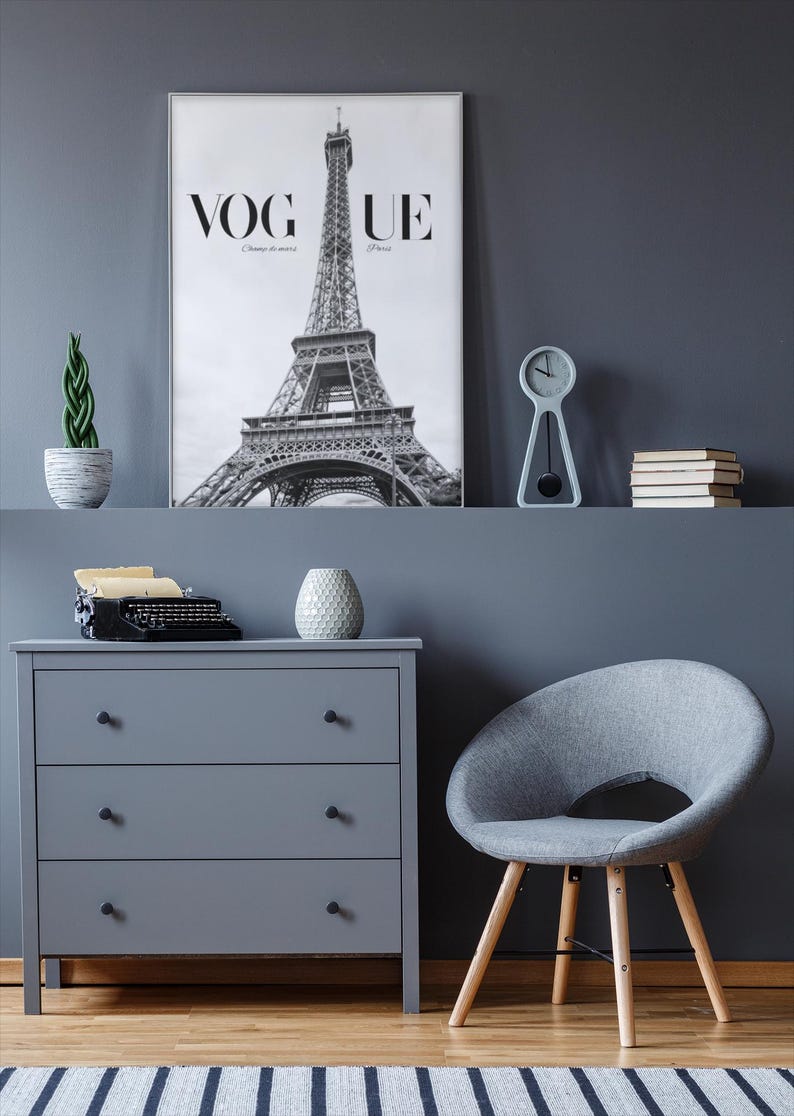 May include: A stylish grey room featuring a Vogue Paris print with the Eiffel Tower. A grey chest of drawers, a grey chair, a small clock, and a potted plant are also present. A vintage typewriter and a decorative vase sit on the chest of drawers.