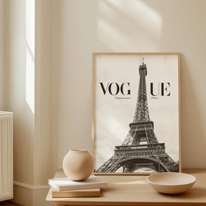 May include: A framed black and white print of the Eiffel Tower with the Vogue logo. The print is on a light wood table with a beige vase, books, and a bowl. The room has a neutral colour palette.