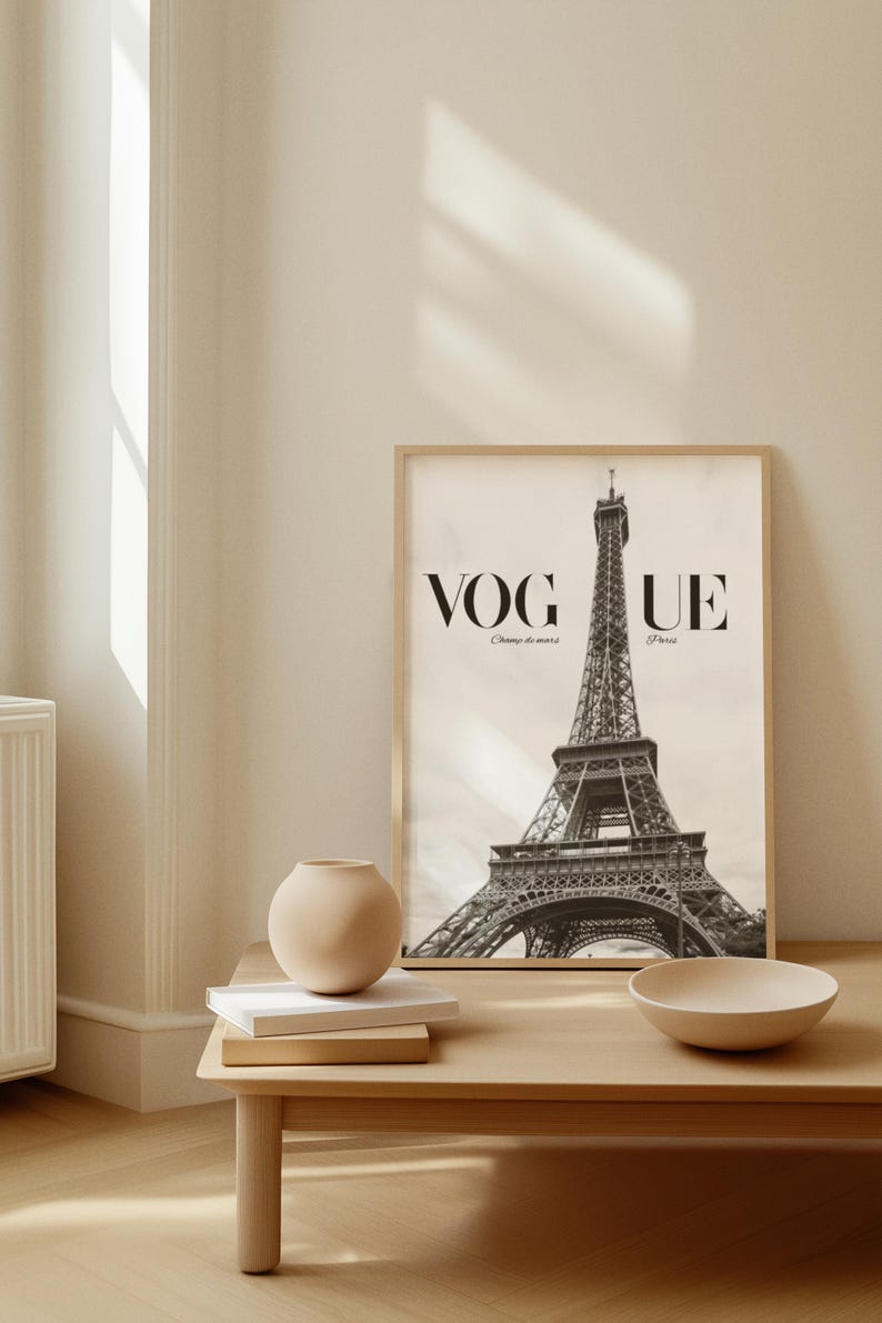 May include: A framed black and white print of the Eiffel Tower with the Vogue logo. The print is on a light wood table with a beige vase, books, and a bowl. The room has a neutral colour palette.