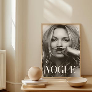 May include: Monochrome framed Vogue magazine cover featuring a woman with a drawn-on moustache. The magazine is on a light wood table with a small vase, books, and a bowl.