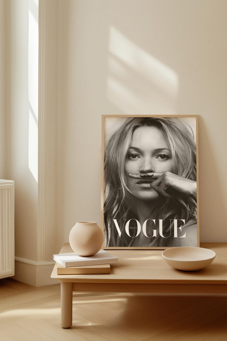 May include: Monochrome framed Vogue magazine cover featuring a woman with a drawn-on moustache. The magazine is on a light wood table with a small vase, books, and a bowl.
