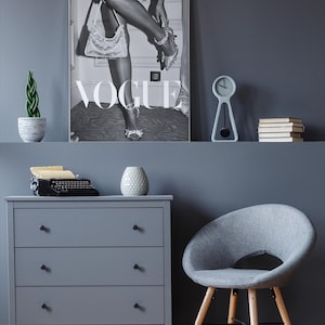 May include: A grayscale Vogue magazine cover print featuring a woman adjusting her shoe, with the Vogue logo prominently displayed. The image is part of a room decor setup, including a gray dresser, chair, and a small clock.