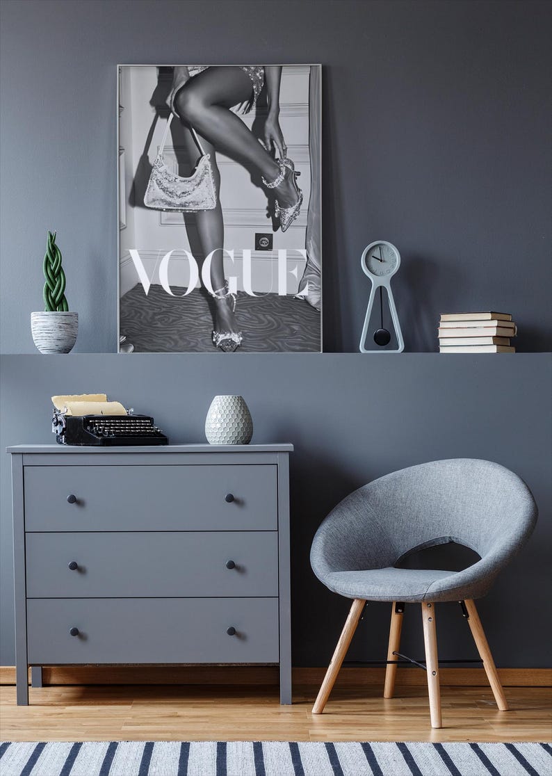 May include: A grayscale Vogue magazine cover print featuring a woman adjusting her shoe, with the Vogue logo prominently displayed. The image is part of a room decor setup, including a gray dresser, chair, and a small clock.