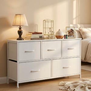 Peut inclure: Une commode blanche à cinq tiroirs avec un cadre en métal. Une lampe, des livres, un présentoir à bijoux, un diffuseur et une photo encadrée sont posés dessus. La commode est dans une chambre.