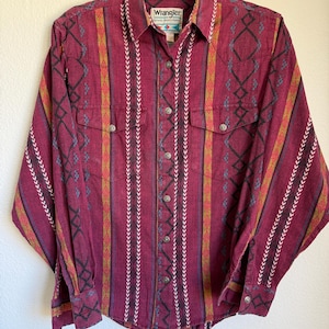 May include: A burgundy button-up shirt with a Western-style design. The shirt features a mix of vertical and geometric patterns in shades of red, yellow, black, and white. It has two chest pockets and the "Wrangler" brand label.