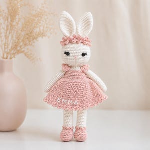 K&ouml;nnte beinhalten: Eine handgefertigte H&auml;kelkaninchenpuppe mit wei&szlig;en und rosa Details. Das Kaninchen tr&auml;gt ein rosa Kleid mit dem Namen "EMMA" darauf, ein passendes Blumenstirnband und rosa Schuhe. Die Puppe hat schwarze Wimpern und eine rosa Nase.
