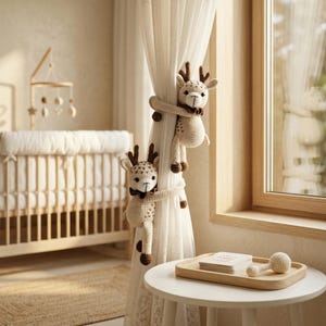 May include: Two cream and brown crocheted deer-shaped curtain tiebacks featuring long arms that wrap around a sheer white curtain. Each deer has embroidered facial features, small antlers, and a dark brown bow tie around its neck.